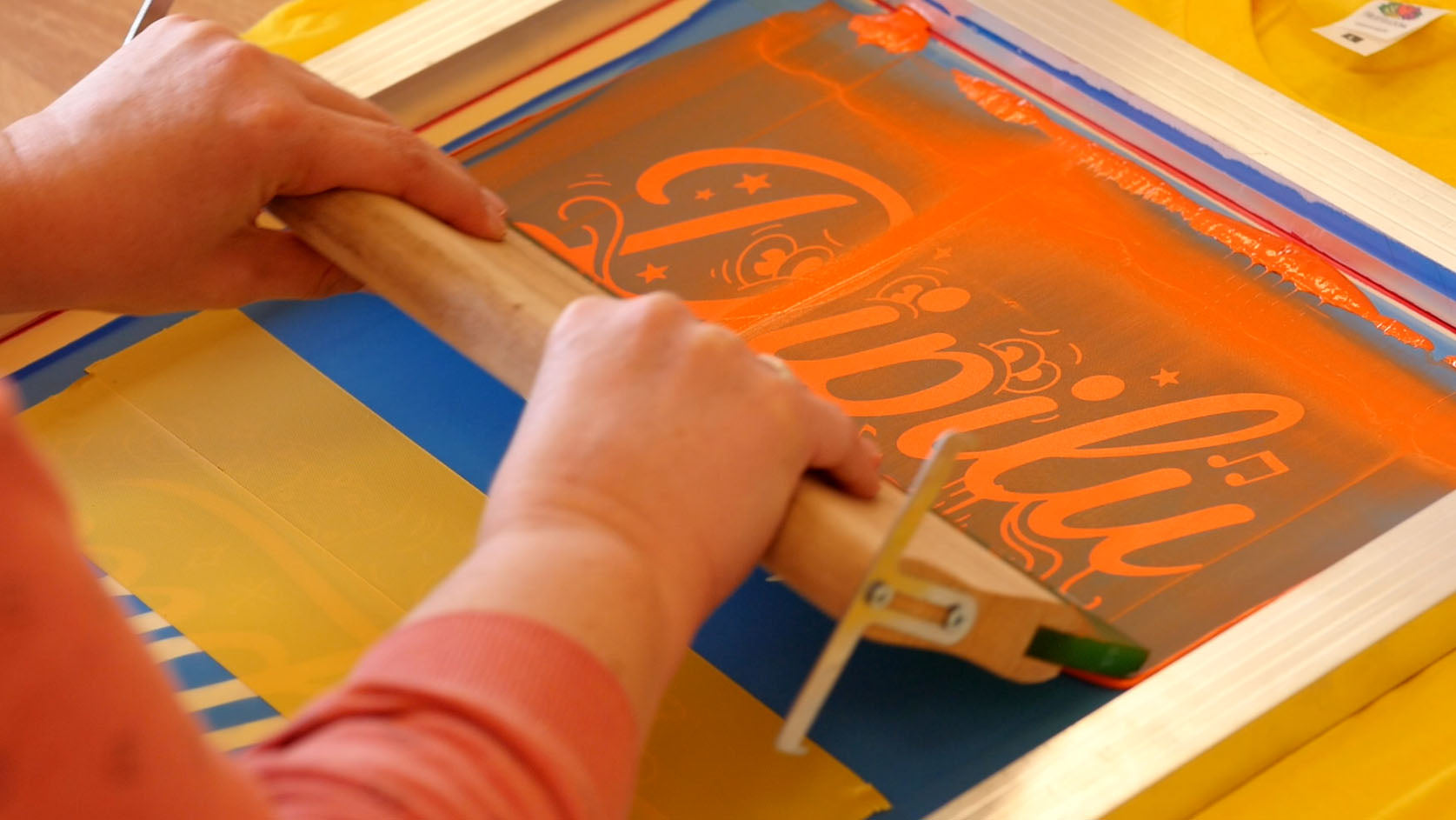 Zwei Hände drucken auf Textil mit einem Siebdruckrahmen, einem Rakel und organener Farbe.