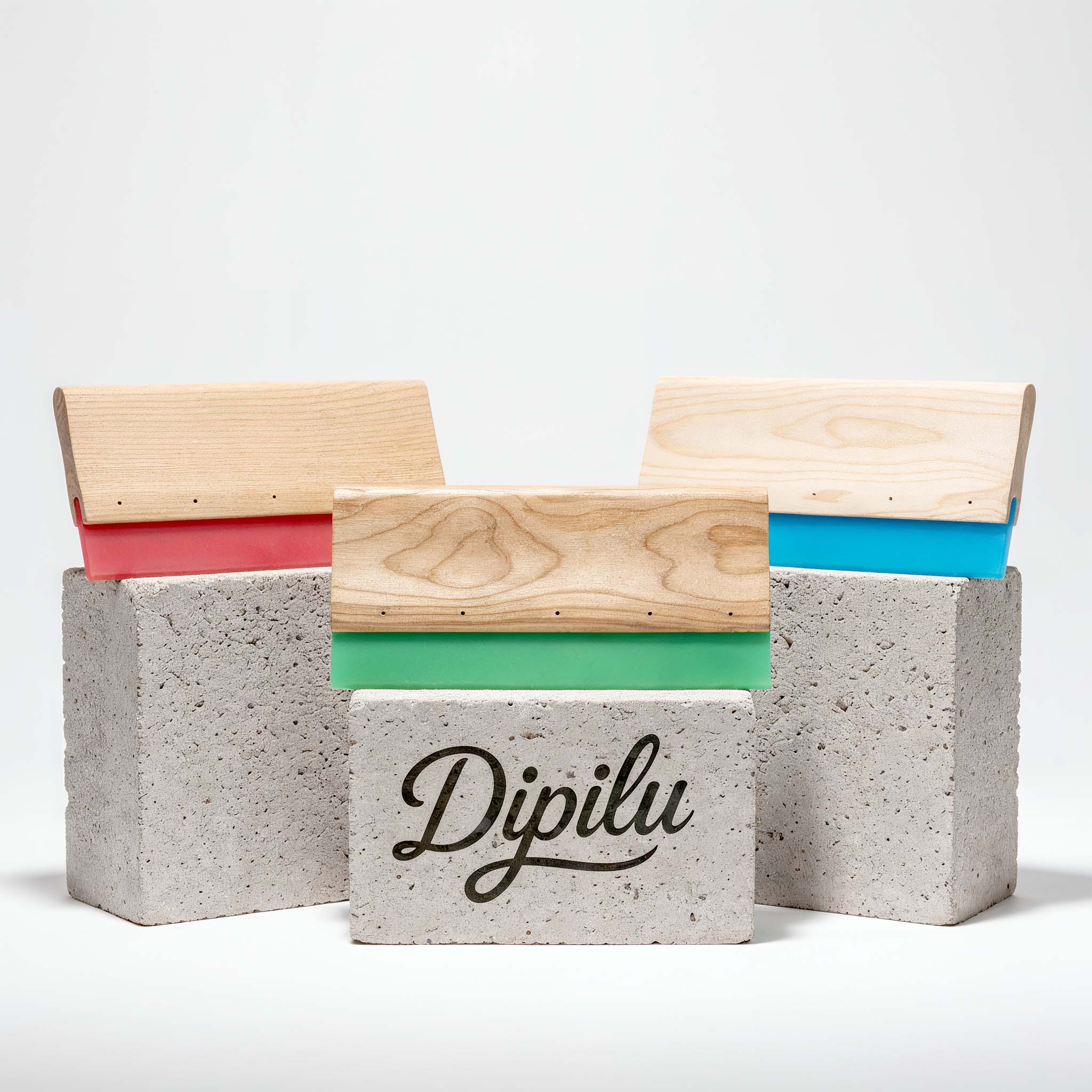 Siebdruckrakel aus Holz mit Gummis unterschiedlicher Farben auf Betonsteinen, der vorderste ist mit dem Dipilu Logo bedruckt.