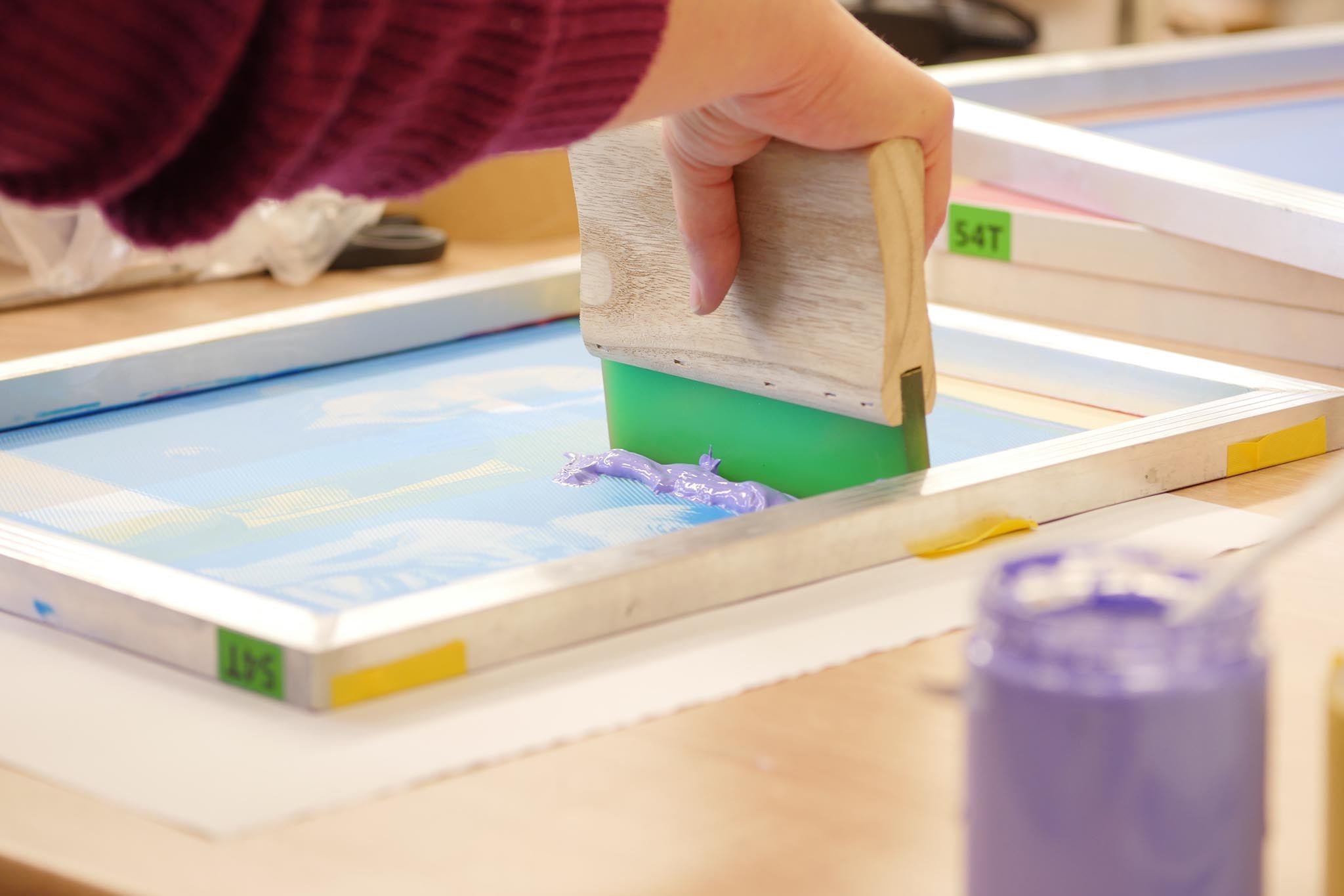 Siebdruckrakel gleitet mit lila Siebdruckfarbe über ein Motiv in einem Siebdruckrahmen