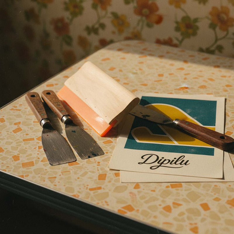 Farbspatel, ein Rakel und Papier mit dem Dipilu Logo bedruckt liegen auf einem Tisch.